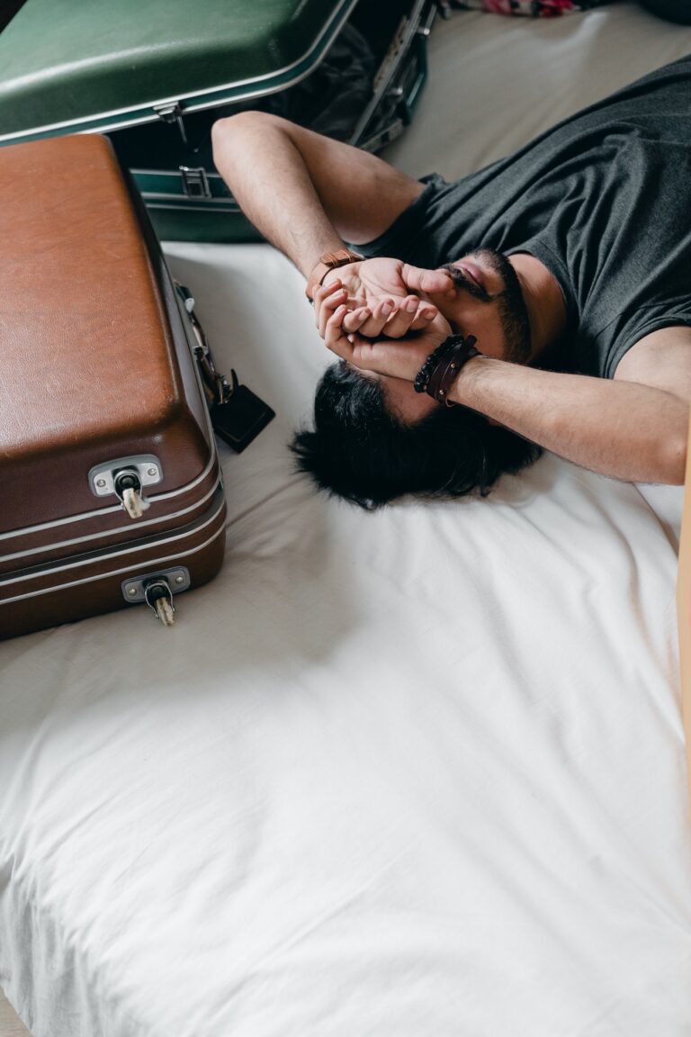 Man with hands over eyes beside suitcases on a bed.