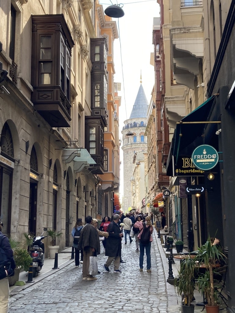 View of Galata Tower from down a cobblestone street lined with restaurants and shops.