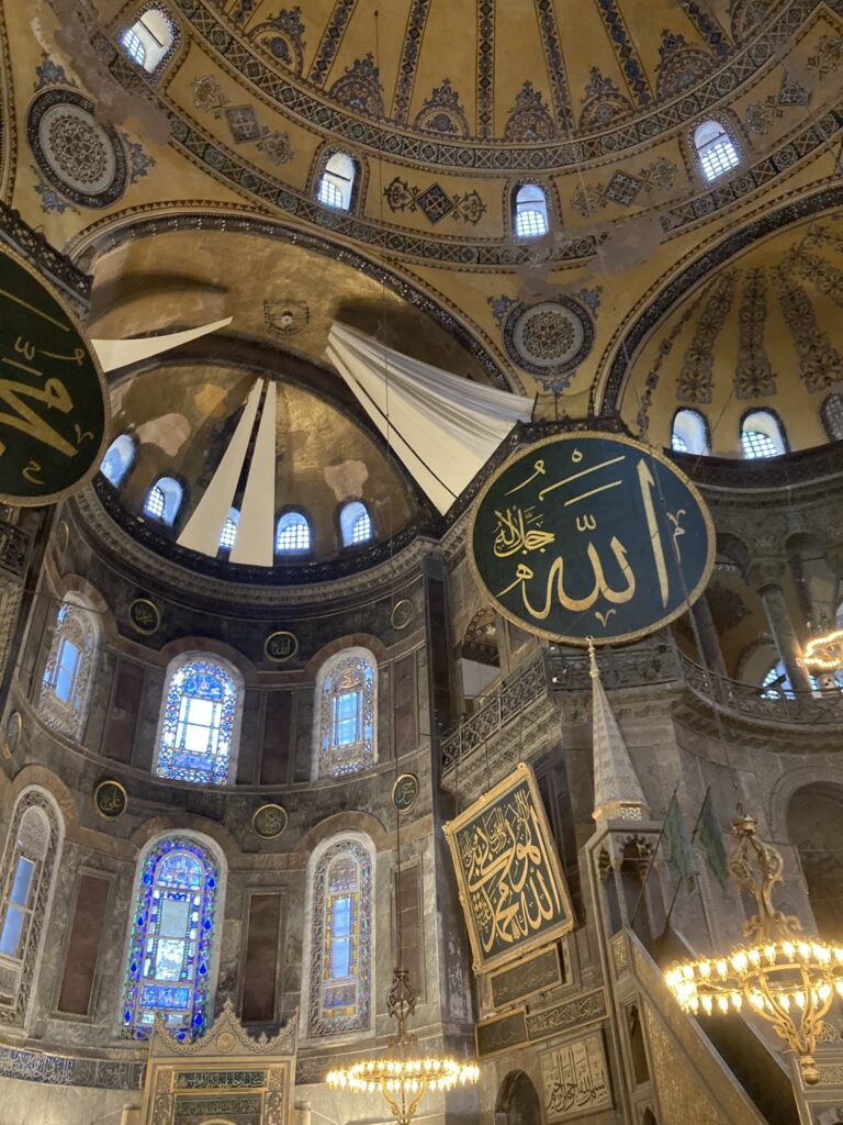 view of dome-shaped roof, arched stained glass windoes, and golden chandeliers from inside Hagia Sophia