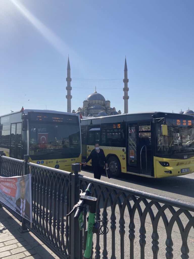 Fenced in sidewalk in Istanbul with two yellow and black public buses stopped in front.