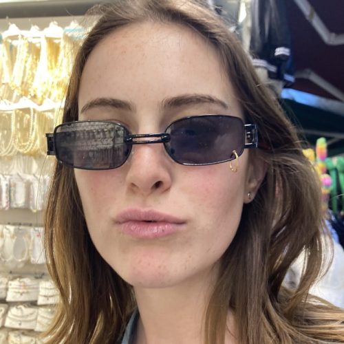 Girl posing with black sunglasses that have a gold ring pierced through one lens.